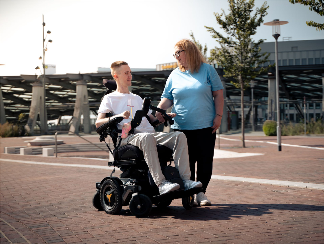 Community care - Patient in wheelchair with caregiver in outdoor public space showing independence and quality of life