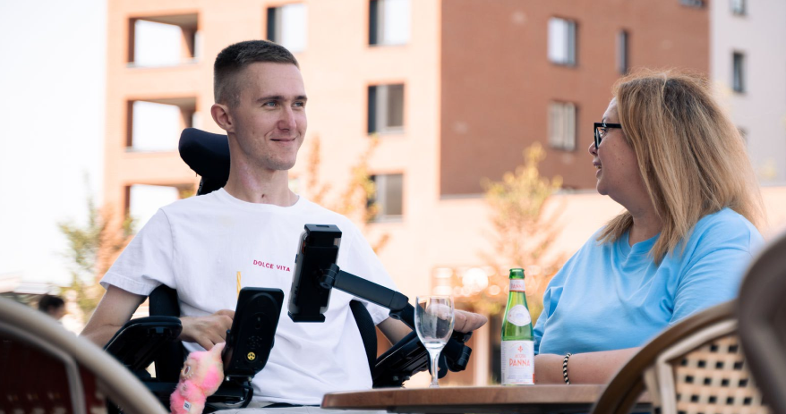 Lebensqualität durch Intensivpflege - Patient im Rollstuhl genießt Zeit mit Betreuerin in gemeinschaftlicher Atmosphäre